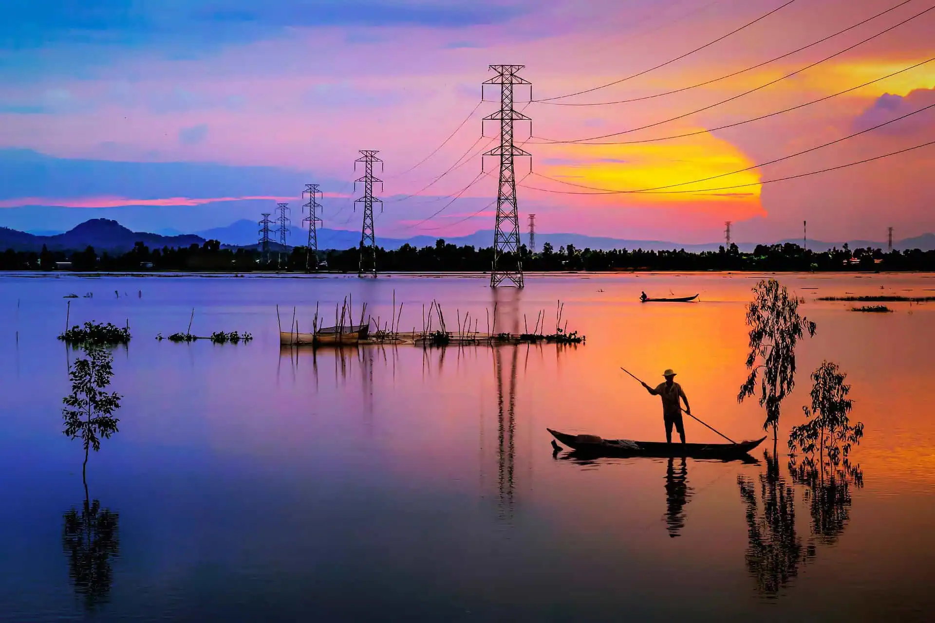 Een sfeervol landschap bij zonsondergang met een uitgestrekte watervlakte waarin hoogspanningsmasten boven de horizon uitsteken. Op de voorgrond is het silhouet van een visser in een bootje zichtbaar, wat het contrast tussen traditionele levenswijzen en moderne energie-infrastructuur benadrukt