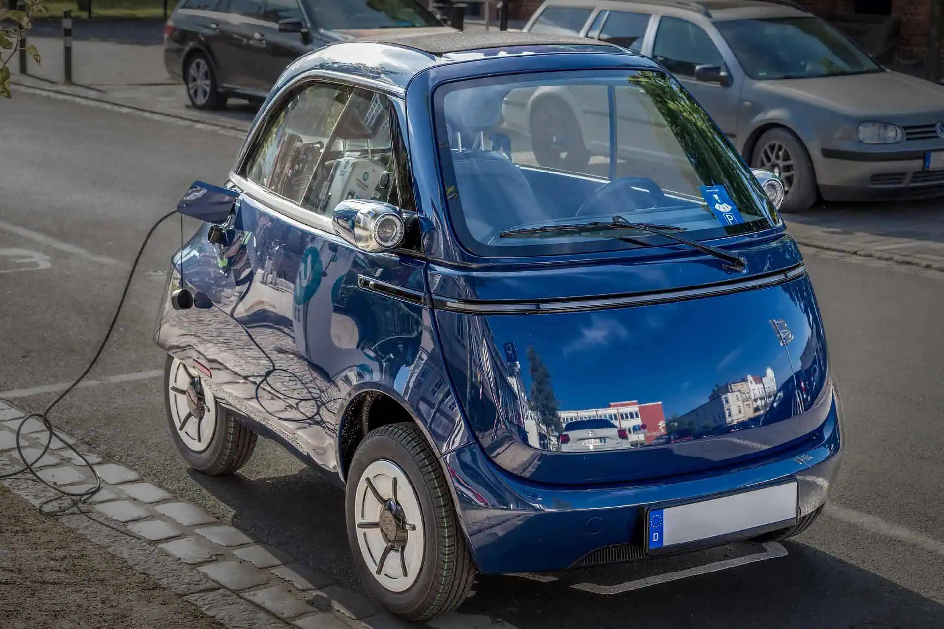 en compacte, donkerblauwe elektrische Microlino stadsauto die aan een laadpaal staat op een geplaveide straat. Het voertuig heeft een retro-ontwerp gebaseerd op de klassieke dwergauto, met een opvallende deur aan de voorzijde