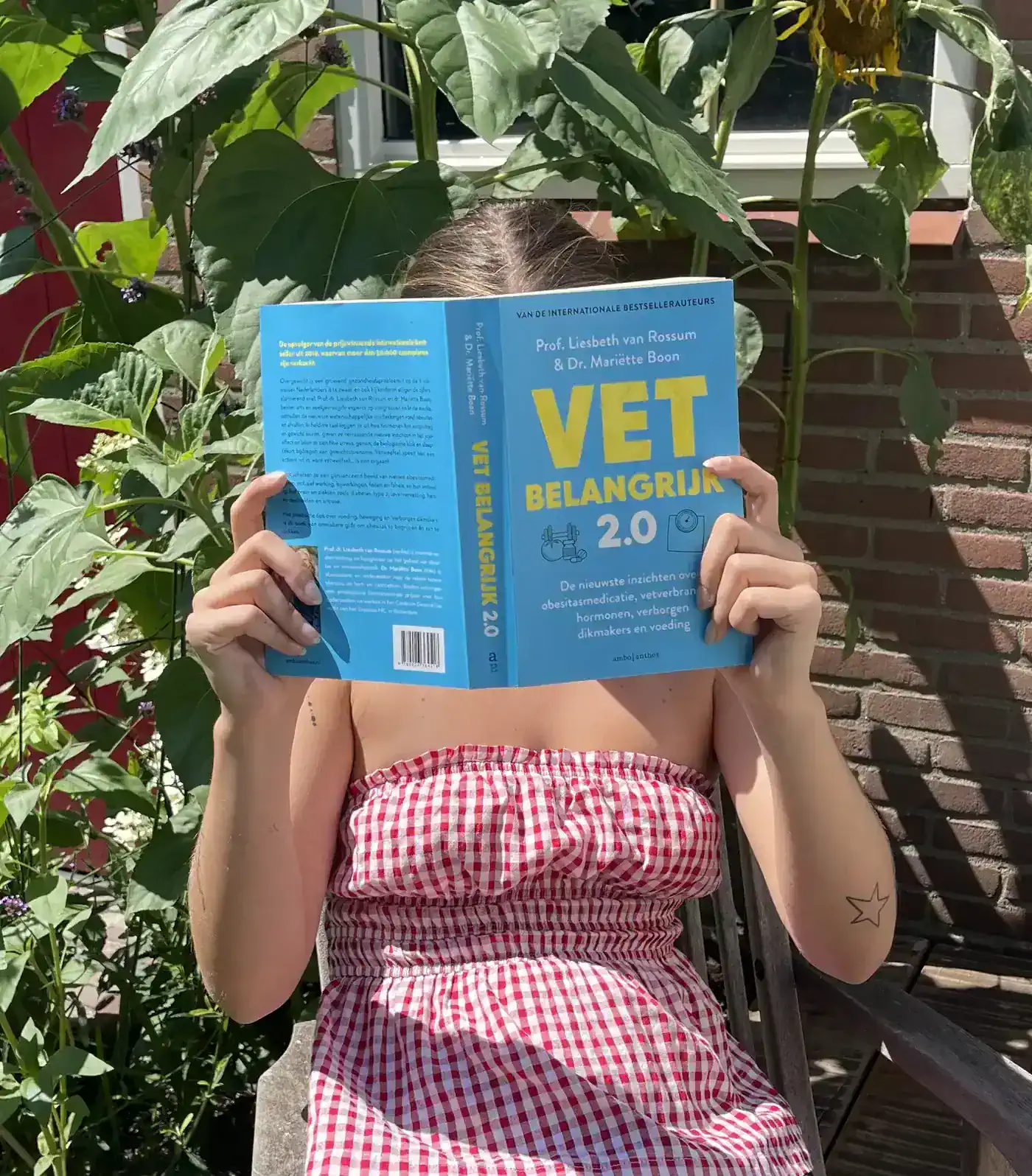 Vrouw in geruite jurk leest het boek Vet belangrijk 2.0 van Liesbeth van Rossum en Mariëtte Boon in een zonnige tuin